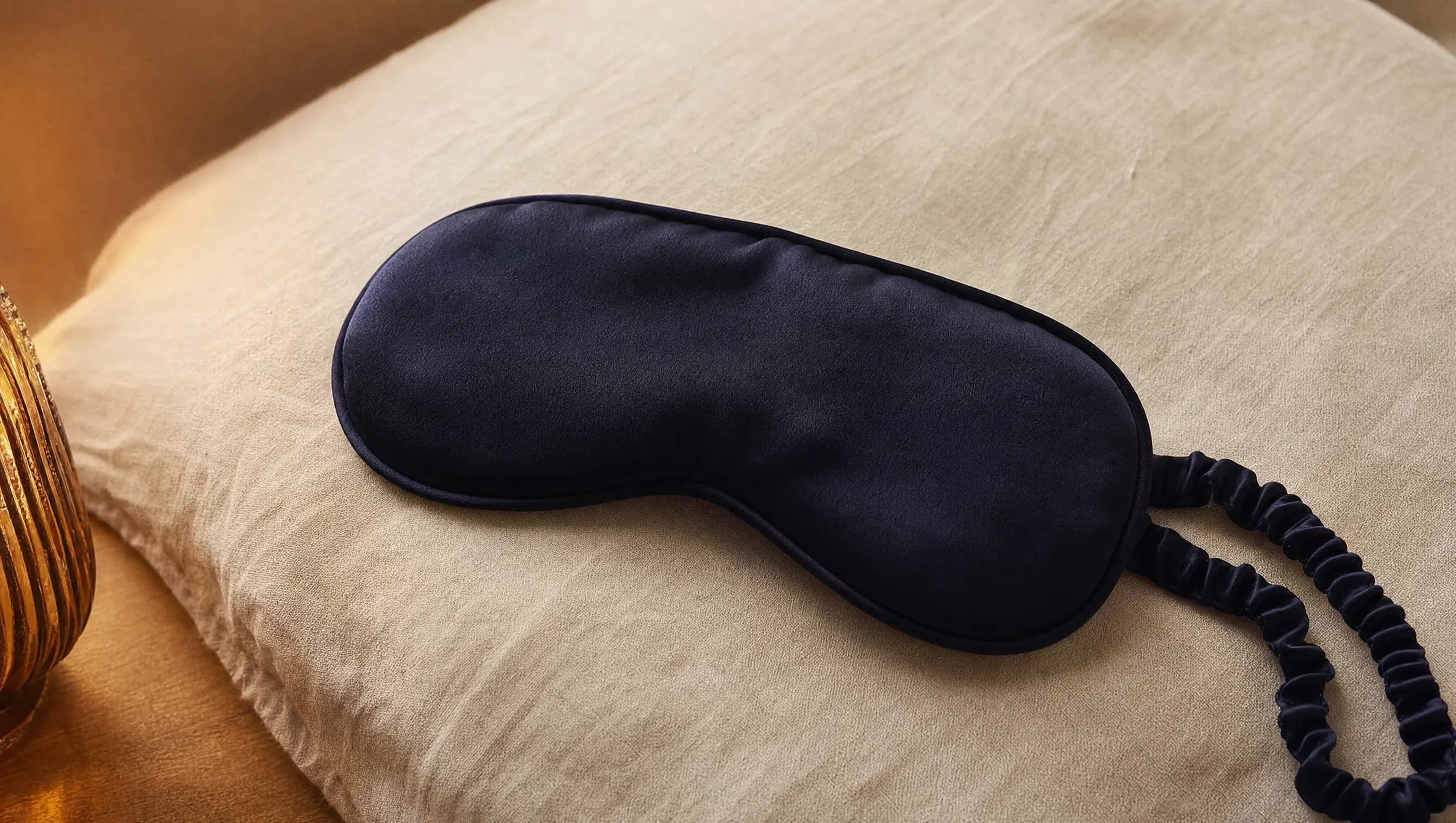 Antifaz de tela oscura doblado sobre una almohada de lino claro con luz suave lateral