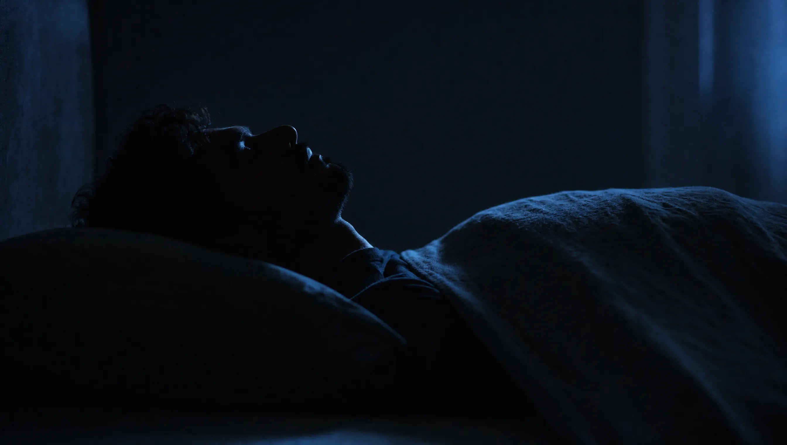 Silueta de una persona dormida boca arriba en penumbra azulada con la cabeza ligeramente inclinada hacia atrás
