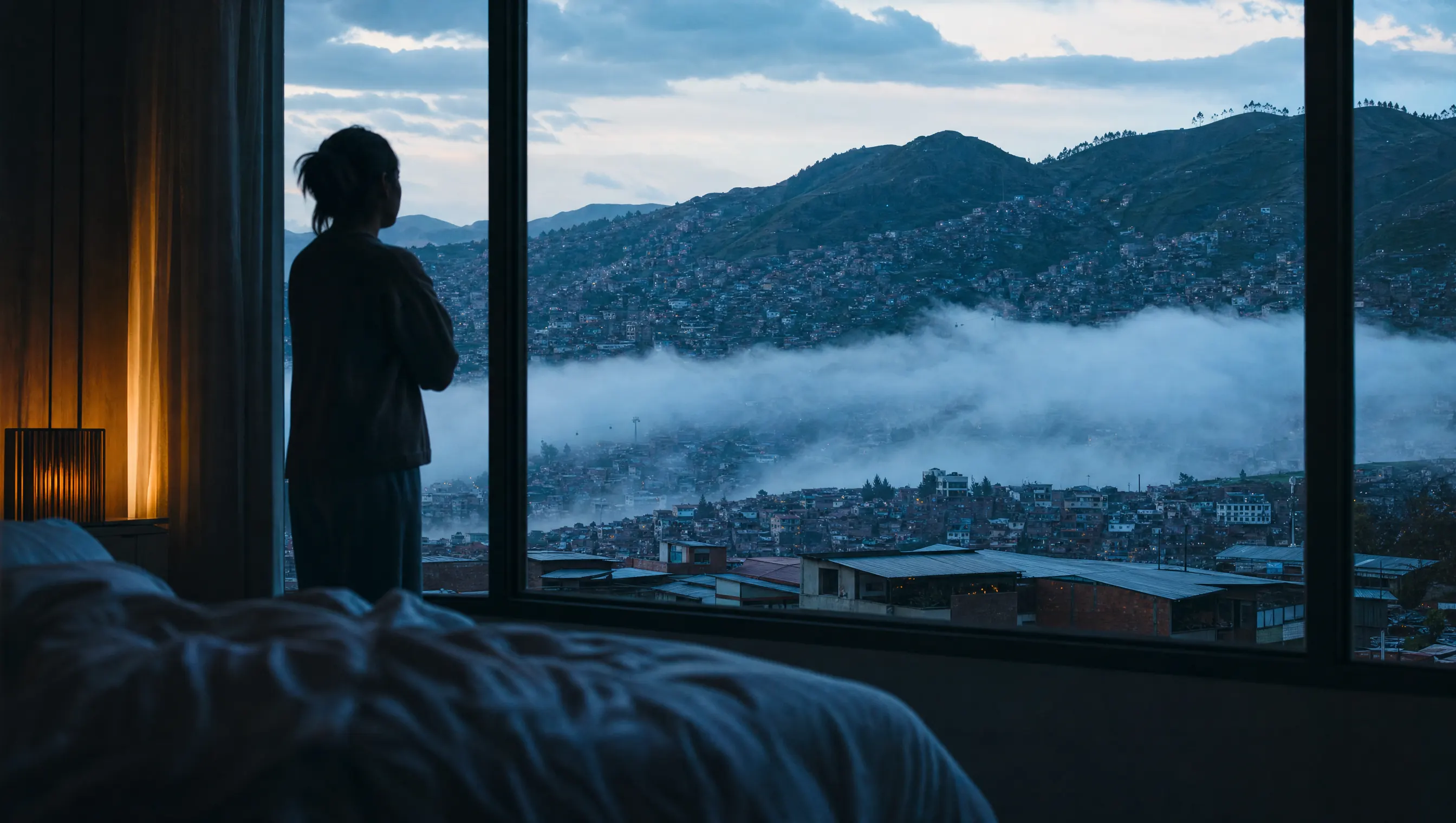 Ventana de dormitorio con vista a una ciudad de montaña al amanecer, con nubes bajas entre los cerros