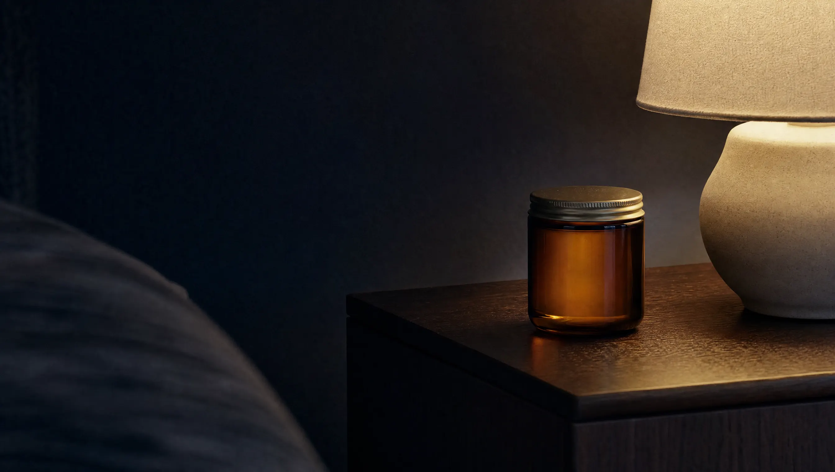 Mesa de noche en penumbra con un frasco de vidrio ámbar cerrado y luz cálida lateral