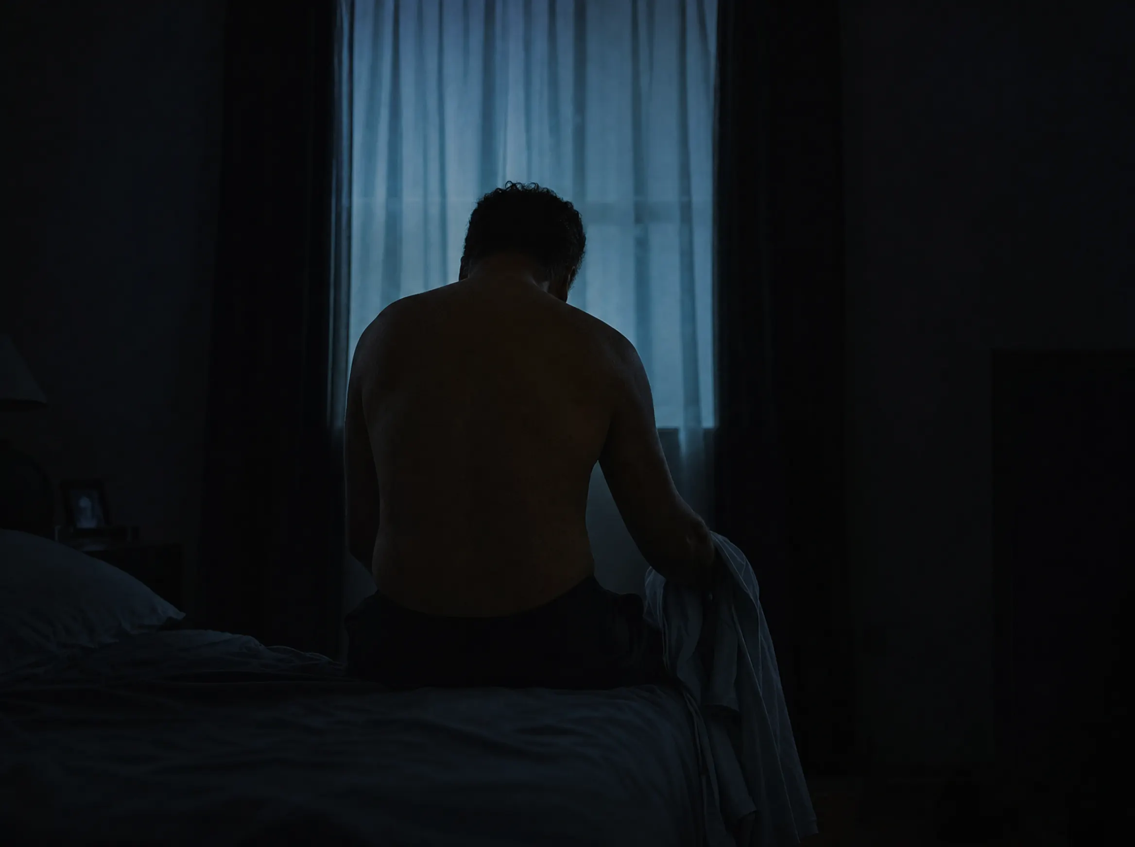 Hombre sentado en el borde de la cama en plena noche, de espaldas, iluminado solo por la luz fría de la ventana